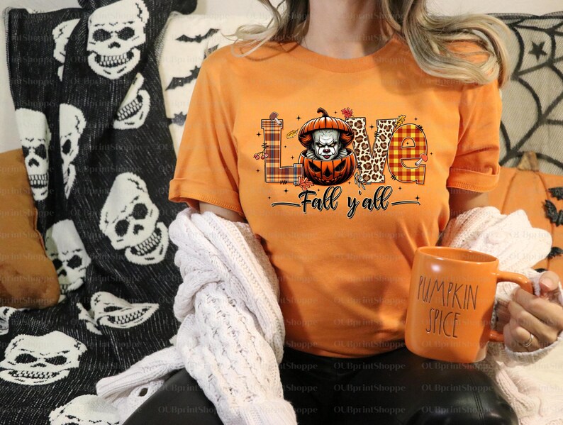 May include: Orange t-shirt with the word "LOVE" in a mix of patterns and a pumpkin with a scary clown face. The text "Fall y'all" is below the word "LOVE". A matching orange mug with "PUMPKIN SPICE" is held in the hand.