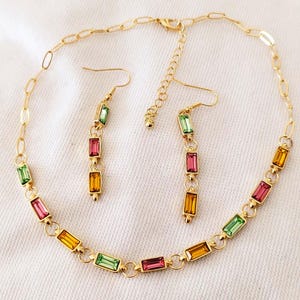Peut inclure: Un ensemble collier et boucles d'oreilles assorties de couleur or. Les bijoux présentent des pierres précieuses rectangulaires et colorées dans les tons vert, rose et jaune. Le collier et les boucles d'oreilles sont présentés sur une surface blanche.