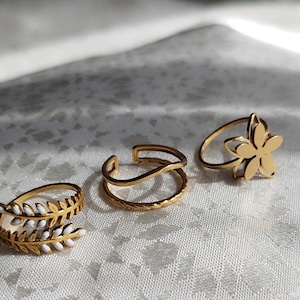Peut inclure: Trois bagues dorées aux designs uniques. L'une présente une double bande, une autre une fleur, et la troisième un motif de feuilles avec des accents d'émail blanc. Les bagues sont présentées sur une surface blanche texturée.