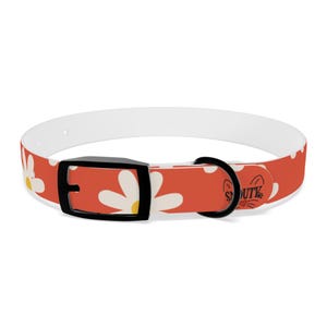 Peut inclure: Un collier pour chien avec un intérieur blanc et un extérieur rouge orné d'un motif de marguerites. Le collier est doté d'une boucle noire et d'un anneau métallique noir. Le mot "SOUTY" est imprimé sur le collier.