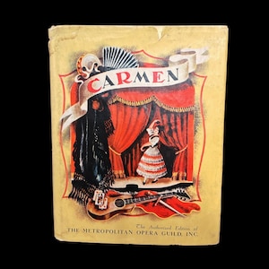 Pode incluir: Capa de livro vintage para "Carmen" com fundo dourado e detalhes em vermelho. A capa apresenta uma cena de palco com uma dançarina, uma guitarra e o texto "CARMEN" em uma faixa. O texto "The Authorized Edition of THE METROPOLITAN OPERA GUILD, INC." também é visível.