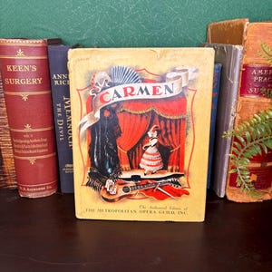 May include: A hardback book titled Carmen, The Authorized Edition of The Metropolitan Opera Guild, Inc. The cover features a yellow background with a central illustration of a dancer on a stage, a guitar, and a fan. The book is surrounded by other books.