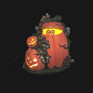 May include: A lighted Halloween decoration featuring three orange pumpkins with carved faces. The largest pumpkin has a pair of eyes visible. Dark green vines and leaves surround the pumpkins, and a power cord extends from the base.
