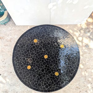 May include: A black round table with a mosaic tile top. The tiles are arranged in a geometric pattern and have a glossy finish. The table has four gold stars in the center.