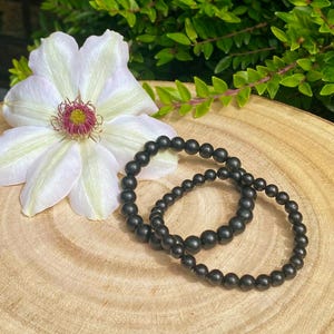 May include: Two black beaded bracelets sit on a wooden surface beside a large white flower with a pink and yellow centre. Green foliage is in the background. The bracelets are made of small, round beads.