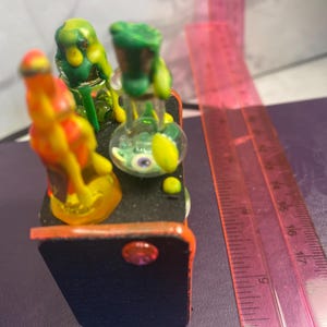 以下が含まれることがあります： 小さな、カラフルなガラス彫刻。オレンジ色の縁取りのある小さな黒い長方形のベースが特徴です。彫刻には、黄色とオレンジ色のフィギュア、緑と黄色のフィギュア、目のデザインのガラスバイアルが含まれています。ピンクの定規が背景にあります。