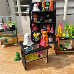 以下が含まれることがあります： 黒い棚と小さなテーブルを備えた、ハロウィーンをテーマにしたミニチュアディスプレイ。 棚には、幽霊のフィギュア、カラフルなポーションボトル、その他の不気味なアクセサリーが飾られています。 配色はオレンジ、緑、ピンクです。