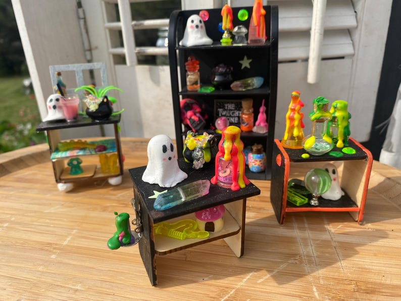 以下が含まれることがあります： 黒い棚と小さなテーブルを備えた、ハロウィーンをテーマにしたミニチュアディスプレイ。 棚には、幽霊のフィギュア、カラフルなポーションボトル、その他の不気味なアクセサリーが飾られています。 配色はオレンジ、緑、ピンクです。