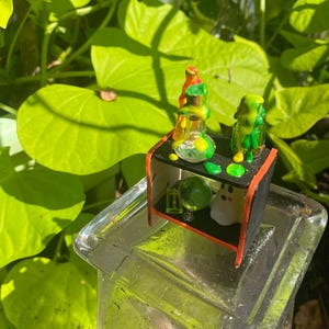 以下が含まれることがあります： ガラスビーカー、緑色のオーブ、小さなゴーストフィギュアを備えた、黒と赤の棚を備えたミニチュアディスプレイ。 棚は透明なガラスの表面に配置され、背景には緑の葉があります。