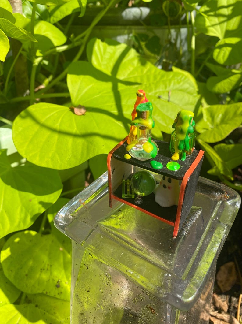 以下が含まれることがあります： ガラスビーカー、緑色のオーブ、小さなゴーストフィギュアを備えた、黒と赤の棚を備えたミニチュアディスプレイ。 棚は透明なガラスの表面に配置され、背景には緑の葉があります。
