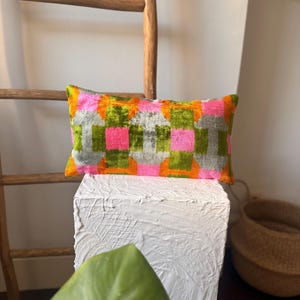 May include: Rectangular decorative pillow with a geometric pattern in shades of pink, green, orange, and gray. The pillow is placed on a white pedestal, with a wooden ladder and a woven basket in the background. The pillow's design features a checkered pattern.
