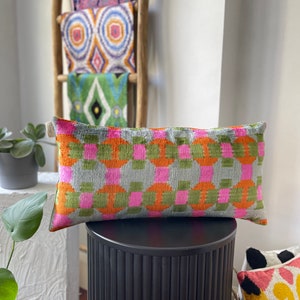 Peut inclure: Un coussin rectangulaire avec un motif géométrique dans des tons de rose, orange, vert et gris. Le coussin est posé sur un piédestal noir.