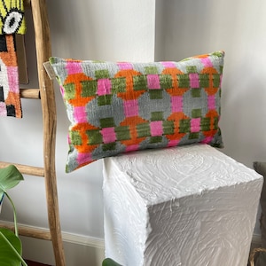 Op de afbeelding: Een rechthoekig kussen met een geometrisch patroon in tinten roze, oranje en groen op een grijze achtergrond. Het kussen ligt op een wit, getextureerd oppervlak.