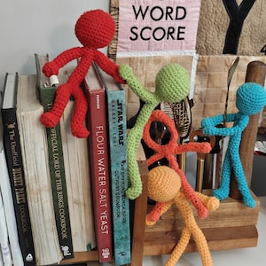 May include: A collection of colorful, crocheted stick figures in red, green, orange, and blue. They are arranged on a wooden shelf with books, including cookbooks and a Lord of the Rings book. A sign above reads "DOUBLE WORD SCORE."