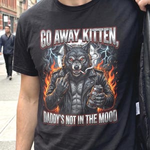 Puede incluir: Camiseta negra con un lobo en chaqueta de cuero, fumando y sosteniendo una lata de cerveza. El texto en la camiseta dice "GO AWAY KITTEN, DADDY'S NOT IN THE MOOD."