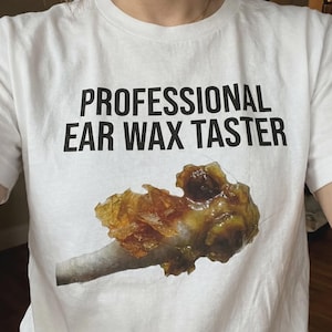 May include: A white short-sleeved t-shirt featuring the black text "PROFESSIONAL EAR WAX TASTER" printed on the chest. Below the text is a realistic graphic illustration of a large, textured, amber and brown-colored mass of ear wax.
