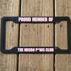 Puede incluir: Un marco de matrícula rectangular negro con texto blanco y borde rosa. La parte superior dice PROUD MEMBER OF y la parte inferior dice THE MICRO P*NIS CLUB. El marco cuenta con cuatro orificios de montaje para fijarlo a un vehículo.