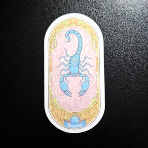 Puede incluir: Pegatina ovalada con una ilustraci&oacute;n de escorpi&oacute;n azul claro sobre un fondo rosa con un patr&oacute;n de remolinos. El dise&ntilde;o est&aacute; enmarcado por un borde dorado y crema, con la palabra "Scorpio" en la parte inferior. Un art&iacute;culo con tem&aacute;tica zodiacal.