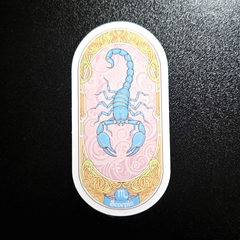Puede incluir: Pegatina ovalada con una ilustraci&oacute;n de escorpi&oacute;n azul claro sobre un fondo rosa con un patr&oacute;n de remolinos. El dise&ntilde;o est&aacute; enmarcado por un borde dorado y crema, con la palabra "Scorpio" en la parte inferior. Un art&iacute;culo con tem&aacute;tica zodiacal.