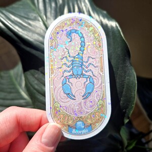 Puede incluir: Una pegatina con una ilustraci&oacute;n de un escorpi&oacute;n azul, el signo zodiacal de Escorpio. El dise&ntilde;o est&aacute; enmarcado en un marco ovalado adornado con un fondo rosa claro y detalles iridiscentes. La palabra "Scorpio" est&aacute; en la parte inferior.