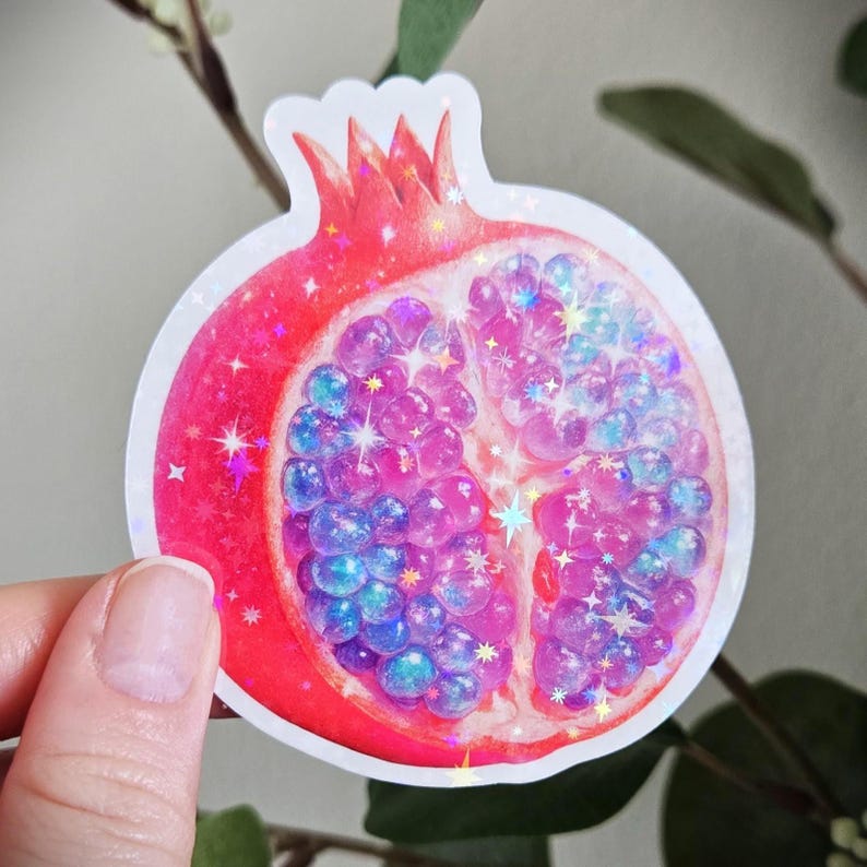 May include: A sticker featuring a stylized pomegranate cut in half. The fruit's exterior is red, while the interior seeds are a mix of pink and blue, with sparkling glitter accents. The sticker has a white border.
