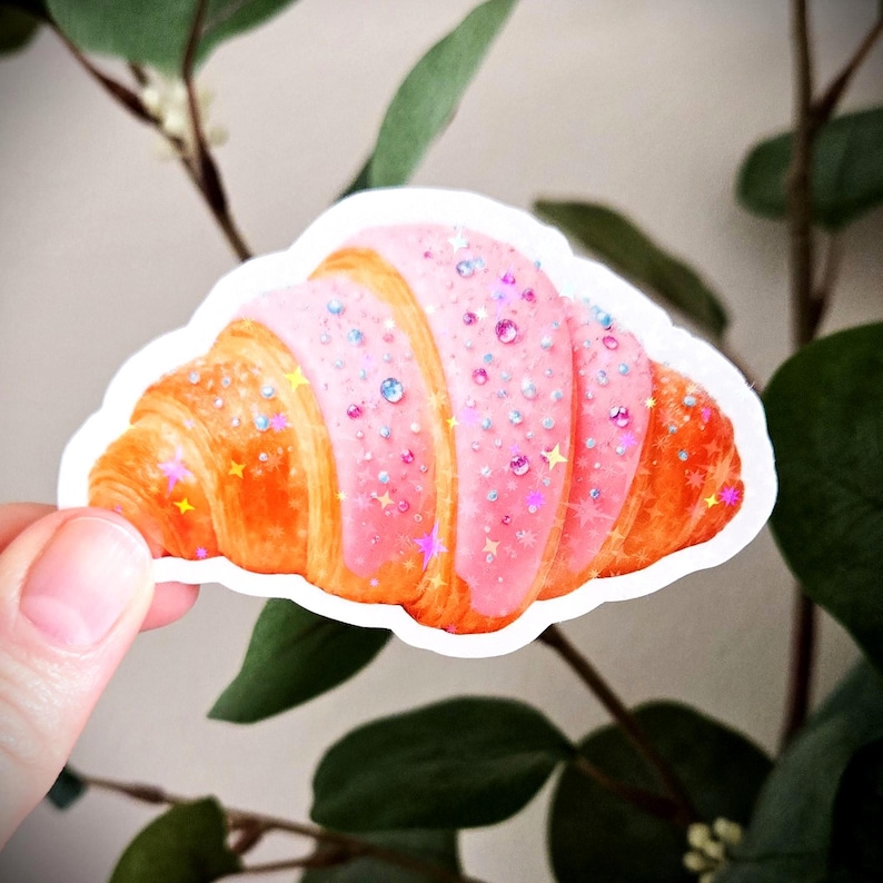 May include: A sticker depicting a croissant with pink icing and glitter. The pastry is a golden brown colour with pink frosting and sparkly stars and gems. The sticker is held in front of a background of green leaves.