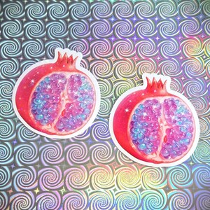 May include: Two pomegranate stickers with a red exterior and a vibrant interior of blue and pink orbs. The stickers are set against a holographic background with a repeating spiral pattern.