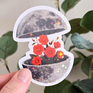 Peut inclure: Un autocollant avec un design céleste. Il représente un bol gris texturé avec des fleurs rouges à l'intérieur, surmonté d'une lune grise. La moitié supérieure de la lune est ouverte, révélant les fleurs. L'autocollant a un effet holographique et scintillant.