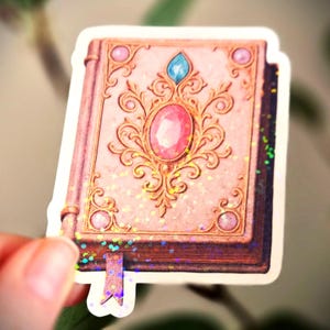 May include: A pink sticker of a book with a detailed gold design on the cover. The cover features a large pink oval gem and a smaller blue gem. The sticker has a glittery effect and a purple ribbon bookmark.
