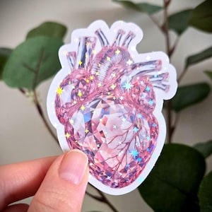 May include: A holographic sticker depicting a stylized human heart, rendered as a gem. The sticker has a pink and iridescent colour scheme with star accents. The design is detailed, with facets and veins visible, creating a unique and eye-catching aesthetic.