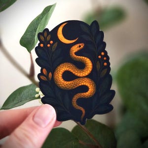 Peut inclure: Un autocollant représentant un serpent doré dans un cadre sombre et orné, avec un croissant de lune et du feuillage. Le design a une ambiance mystique. L'autocollant est tenu devant un fond de feuilles vertes.