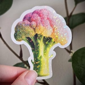 May include: A colorful sticker of a broccoli floret with a rainbow gradient of colors, including green, yellow, pink, and purple. The sticker has a glittery finish and a white outline. The sticker is held by a person's hand.