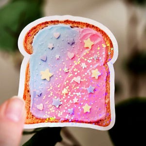 May include: A sticker featuring a slice of toast with a gradient of blue and pink frosting. The frosting is decorated with star and heart-shaped sprinkles and glitter. The toast has a brown crust and is held by a hand.