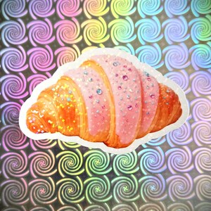 May include: A sticker depicting a croissant with pink icing and colourful sprinkles. The pastry is golden brown. The background features a pattern of swirling rainbow-coloured shapes.