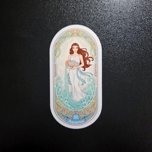 Pu&ograve; includere: Un adesivo ovale raffigurante il segno zodiacale dell'Acquario. Raffigura una donna con lunghi capelli rossi, che indossa un abito bianco, che tiene in mano un'anfora. Il design include elementi d'acqua blu e turchese e bordi decorativi.