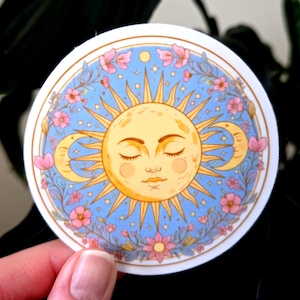 May include: A circular sticker with a celestial theme. It depicts a yellow sun with closed eyes, accompanied by crescent moons, on a light blue backdrop. Pink flowers and stars encircle the sun and moons, creating a decorative and whimsical design.