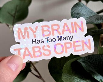 以下が含まれることがあります： ピンクと黒で「MY BRAIN Has Too Many TABS OPEN」と書かれた白いステッカー。ステッカーはホログラフィックでキラキラした効果があります。ステッカーは人の手で持たれており、背景には緑の葉があります。