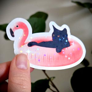 May include: A sticker depicting a black cat lounging on a pink flamingo inflatable. The cat has glittery details and is set against a white background. The sticker has a holographic effect with star and glitter accents.