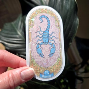 Puede incluir: Una pegatina con una ilustraci&oacute;n de un escorpi&oacute;n azul claro, el signo zodiacal de Escorpio. El dise&ntilde;o est&aacute; sobre un fondo rosa con un borde decorativo. La palabra "Escorpio" est&aacute; escrita en una pancarta azul en la parte inferior.