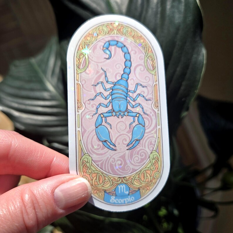 Puede incluir: Una pegatina con una ilustraci&oacute;n de un escorpi&oacute;n azul claro, el signo zodiacal de Escorpio. El dise&ntilde;o est&aacute; sobre un fondo rosa con un borde decorativo. La palabra "Escorpio" est&aacute; escrita en una pancarta azul en la parte inferior.