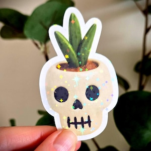 May include: A sticker of a skull-shaped plant pot with a small succulent. The beige pot has black eye sockets and a mouth. The sticker has a holographic, glittery finish with star-shaped sparkles.