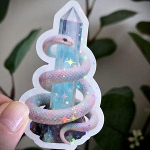 May include: A sticker depicting a pastel-coloured snake coiled around a light blue crystal. The snake has pink, purple, and blue scales. The crystal is decorated with colourful star-shaped accents. The sticker has a white border.