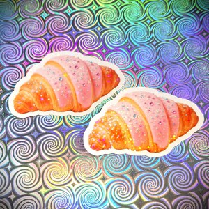 May include: Two stickers of croissants with pink icing and sparkling sprinkles. The croissants are golden-brown against a holographic background with a swirl pattern. Each sticker is approximately 8 cm long.