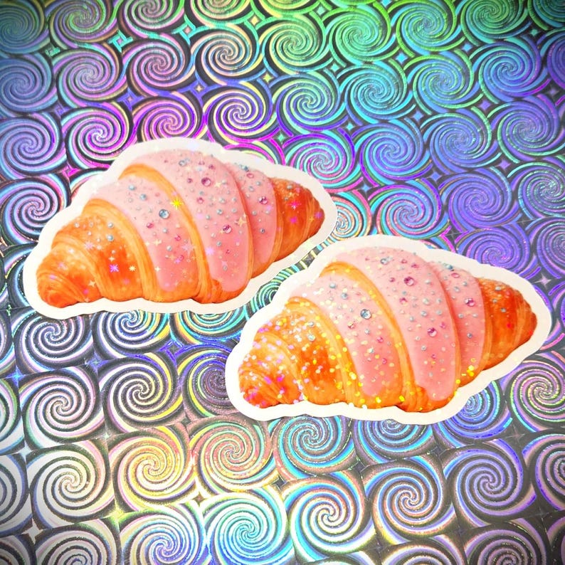 May include: Two stickers of croissants with pink icing and sparkling sprinkles. The croissants are golden-brown against a holographic background with a swirl pattern. Each sticker is approximately 8 cm long.