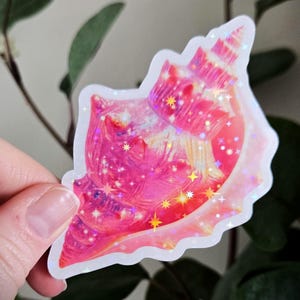 May include: A pink and iridescent seashell sticker with a holographic effect. The sticker features a detailed design of a conch shell with a wavy edge, and is decorated with star-shaped accents.