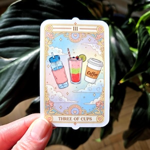May include: A sticker featuring a tarot card design with the text "THREE OF CUPS." The card depicts three beverages: a smoothie, a coffee cup, and a water bottle. The card has a pastel color scheme with clouds and decorative elements.