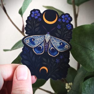 May include: A dark sticker featuring a detailed illustration of a moth with blue and white patterned wings, set against a backdrop of dark foliage, blue flowers, and crescent moons. The moth has intricate designs, and the sticker is held by a hand.