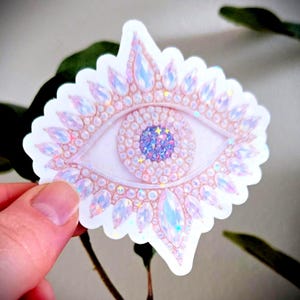 Peut inclure: Autocollant représentant un œil stylisé orné de pierres précieuses irisées roses, bleues et violettes. L'iris est un mélange scintillant de paillettes bleues et violettes. Le bord de l'autocollant est festonné.