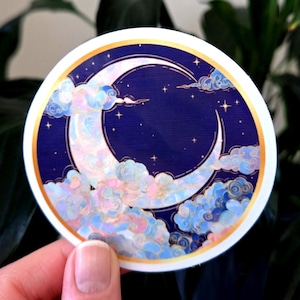 Puede incluir: Pegatina redonda con un diseño de luna creciente sobre un fondo azul oscuro con detalles dorados. Nubes y estrellas en tonos pastel rodean la luna, creando un tema celestial. La pegatina tiene un borde blanco.
