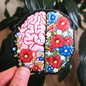 May include: A sticker with an embroidered design of a brain divided into two halves. One side shows a pink brain, the other is filled with red and blue flowers. The sticker has a black border and is held by a hand.